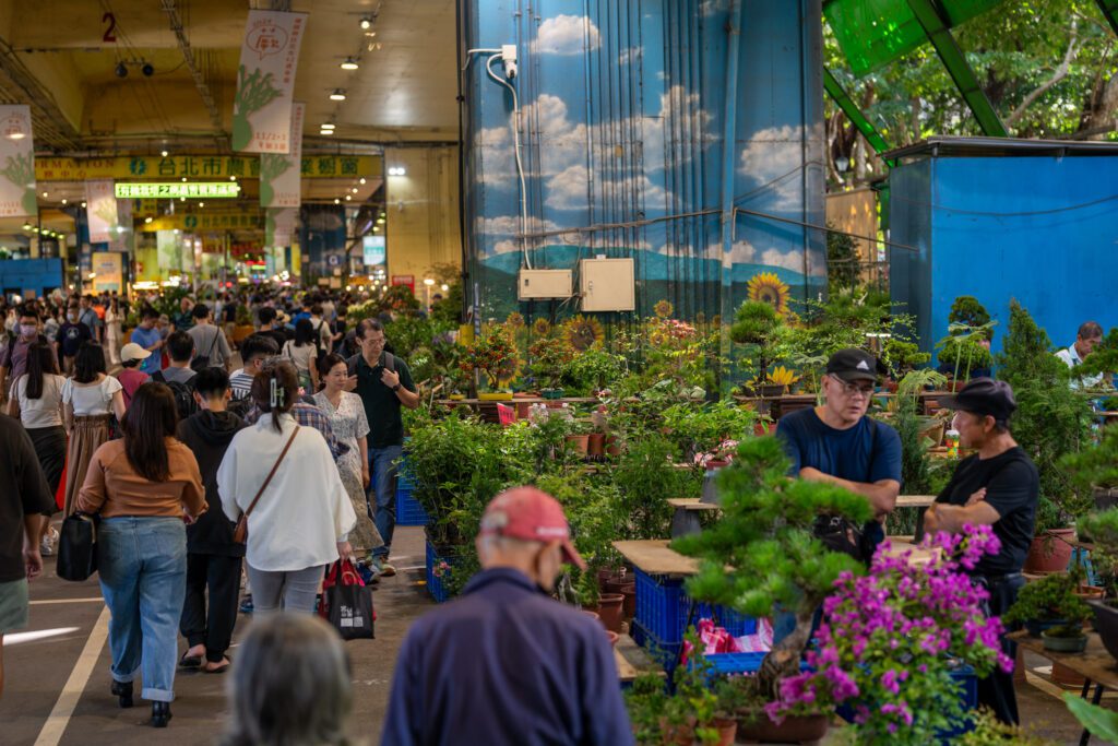 A bustling market scene with people walking and shopping for plants, surrounded by vibrant greenery and colorful flowers, under a canopy.