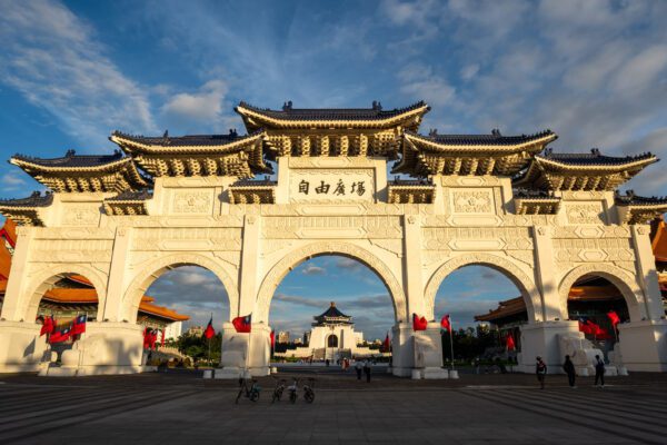 Ornate gateway at Liberty Square in Taipei, bathed in golden light. A few people and red flags are visible around the majestic arches.