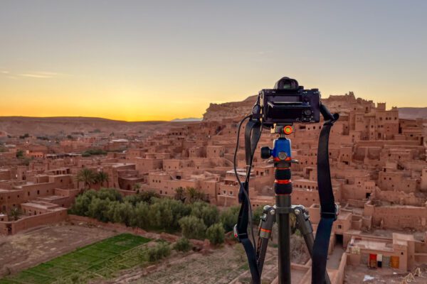 A camera on a tripod overlooks Ait Benhaddou at sunset, capturing the clay architecture and desert landscape of the historic Moroccan site.