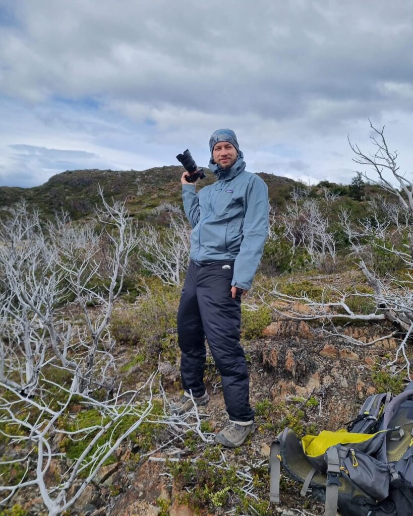 A person holding a camera stands on rocky terrain with sparse vegetation, under a cloudy sky. A backpack is on the ground nearby.