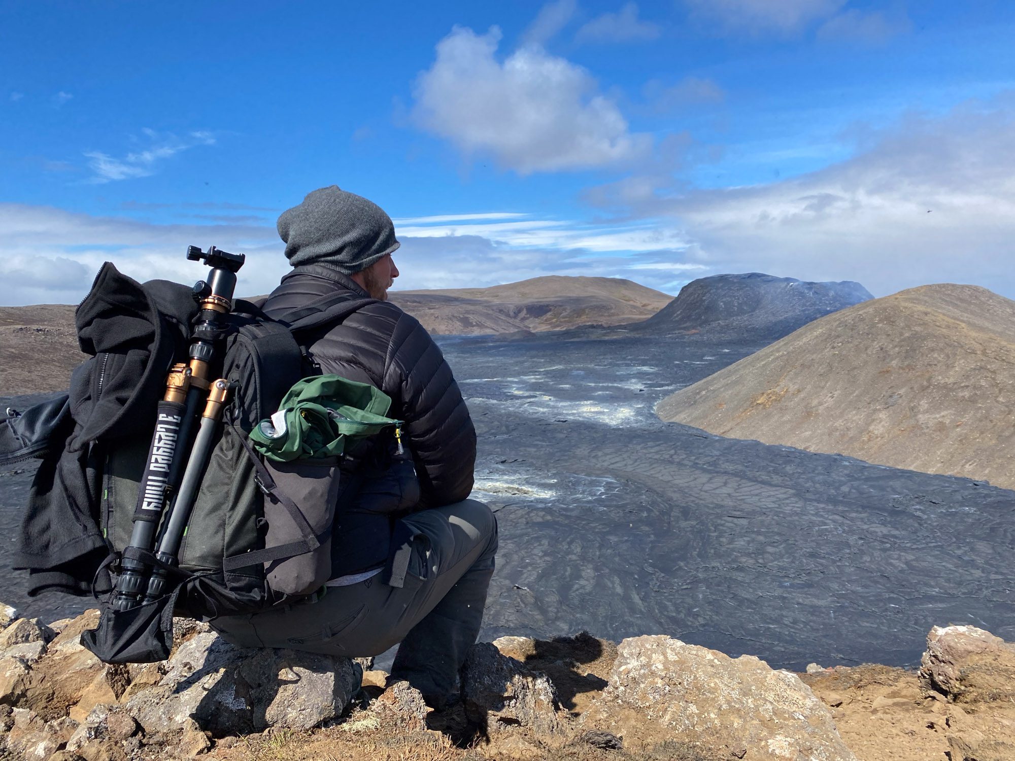 A person with a backpack sits on rocky terrain, overlooking a vast, barren landscape under a bright sky. Hills stretch into the distance.