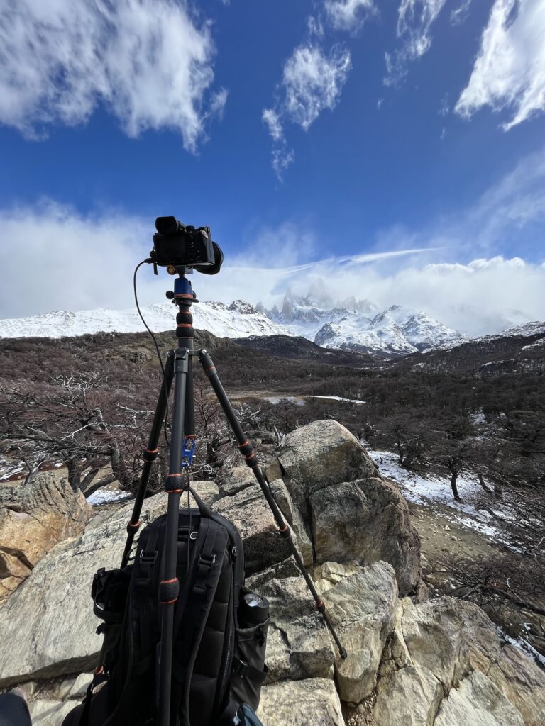 Camera on tripod overlooking snow-capped mountains in Patagonia. Rugged landscape, cloudy sky, and rocky terrain provide a stunning, natural backdrop for photography.